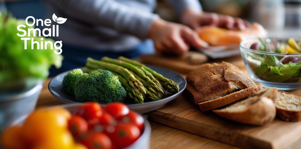 A kitchen counter with fresh food laid out for meal preparation, including a plate of broccoli and asparagus, a bowl of cherry tomatoes, a loaf of sliced bread on a wooden board, and a bowl of mixed salad. In the background, a person is preparing food. The words ‘One Small Thing’ with a small leaf icon appear in the top left corner.