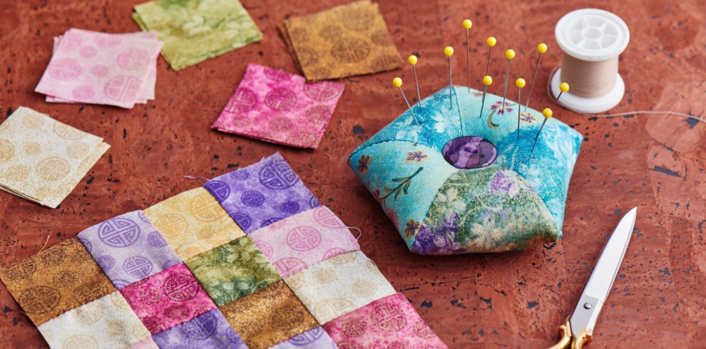 A tabletop scene showing a small quilting project in progress. Several colourful fabric squares in pink, purple, green, gold, and patterned prints are arranged on the surface, with some sewn together into a patchwork piece. Beside them is a handmade, turquoise‑and‑purple fabric pincushion holding several yellow‑topped pins. A spool of white thread and a pair of silver-and-gold scissors sit nearby on the textured brown surface.
