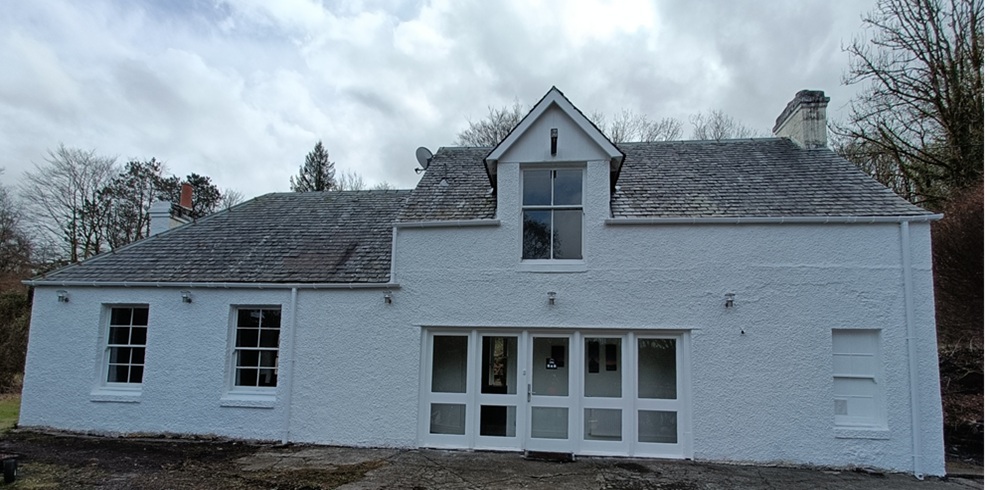 Front view of a white, rough‑cast rural house with a grey slate roof, central gabled dormer window, and multiple white‑framed windows and doors. The building sits on a stone forecourt with trees behind it under an overcast sky.