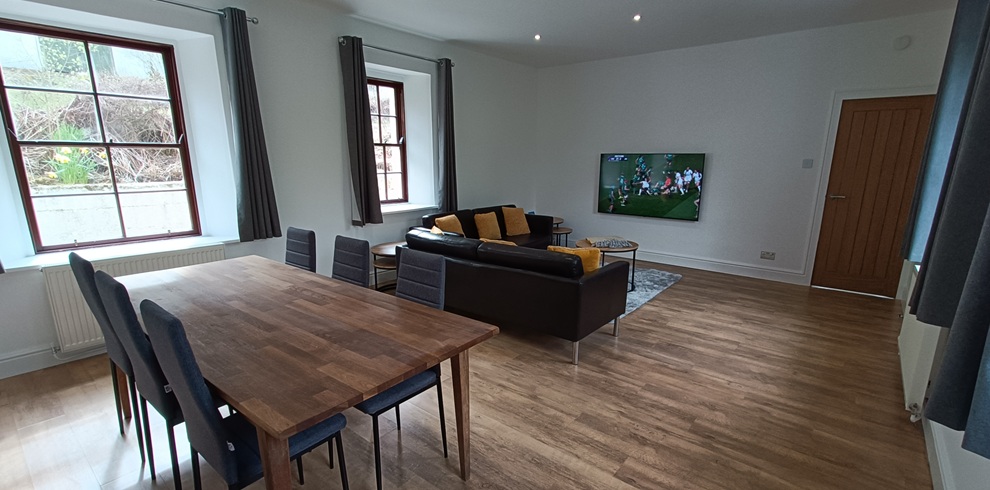 Open‑plan living and dining room with light wood flooring, white walls, and large windows with dark curtains. A wooden dining table with black chairs sits in the foreground, while a black sofa with yellow cushions faces a wall‑mounted television. A wooden door and neutral décor complete the bright, modern space.