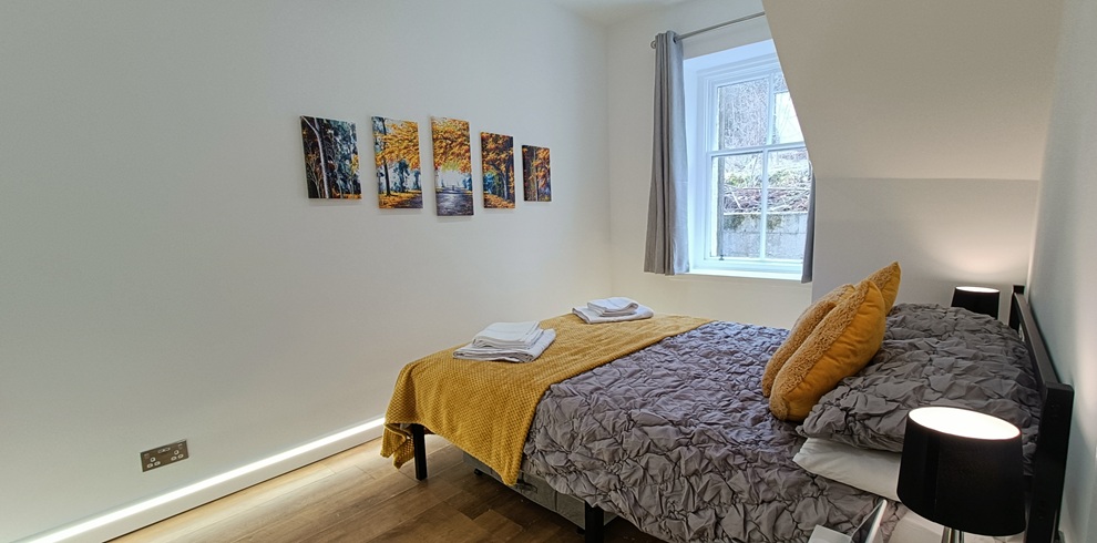 Bright bedroom with white walls and light wood flooring, featuring a double bed dressed in grey bedding and a mustard yellow throw with matching cushions. Folded towels rest on the bed, a small bedside table with a black lamp sits to the right, and a window with grey curtains lets in natural light. A row of framed artwork hangs above the bed.