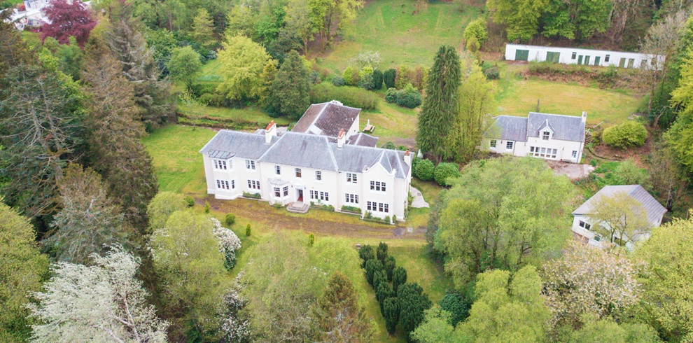 Aerial view of a large white country house surrounded by lush green gardens and mature trees, with several smaller outbuildings nearby, set within a wooded rural landscape.