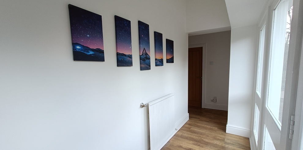 Narrow hallway with white walls and wooden flooring, featuring five framed landscape prints arranged in a row on the left wall above a white radiator, with natural light coming in from a window on the right and a wooden door at the far end.
