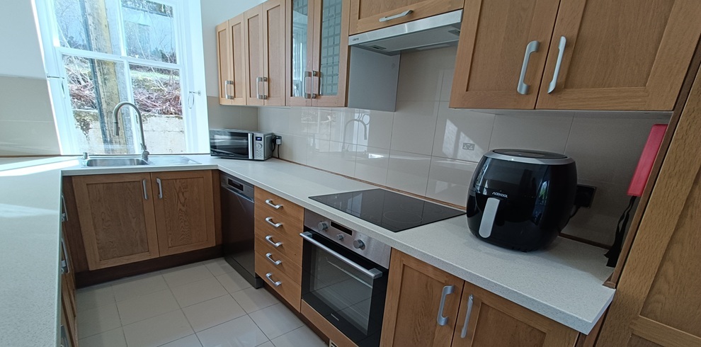 Bright, compact kitchen with wooden cabinets and light countertops, featuring an integrated oven and stovetop, microwave, air fryer, and sink beneath a window, with neutral tiled flooring and walls.