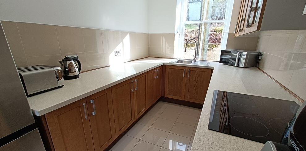 Light-filled kitchen with wooden base cabinets and pale countertops arranged in an L-shape, featuring a ceramic stovetop in the foreground, kettle and toaster on the counter, and a sink beneath a large window with tiled flooring and neutral wall tiles.