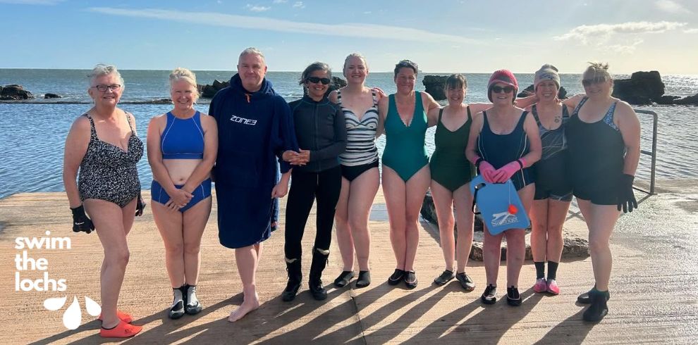 A group of eleven people stand together on a concrete slipway beside the sea, all wearing swimming or outdoor water‑swim clothing such as swimsuits, neoprene boots, gloves, and robes. The sea is behind them, with waves, rocks, and a bright sky with scattered clouds. Sunlight reflects off the water, casting long shadows. On the left side of the image, the words “swim the lochs” appear alongside a small graphic of three water droplets.
