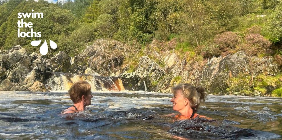 Two people are swimming in an outdoor natural pool surrounded by rocky terrain and dense green vegetation. A small waterfall cascades over the rocks behind them, creating ripples in the water. The swimmers are near the surface, with only their heads and upper shoulders visible above the water. In the top-left corner, the text “swim the lochs” appears beside a simple graphic of three water droplets.