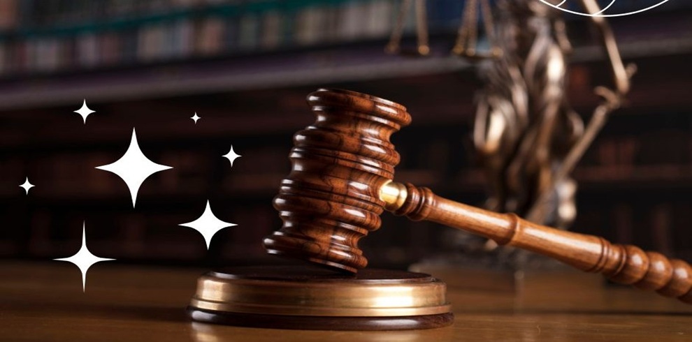 Close-up of a wooden judge’s gavel resting on a sound block on a desk, with shelves of law books blurred in the background and a faint statue of justice visible. White star-like graphic sparkles appear to the left of the gavel.