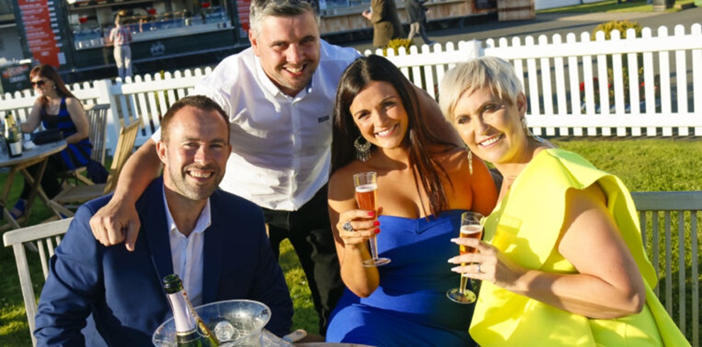 Four adults sit and stand closely together at an outdoor event on a grassy area, holding champagne flutes. One person sits at a table with an ice bucket and bottles, while three others pose nearby. They wear formal or semi-formal clothing in dark blue, white, bright blue, and yellow. A white picket fence, outdoor seating, and event structures are visible in the background under bright sunlight.