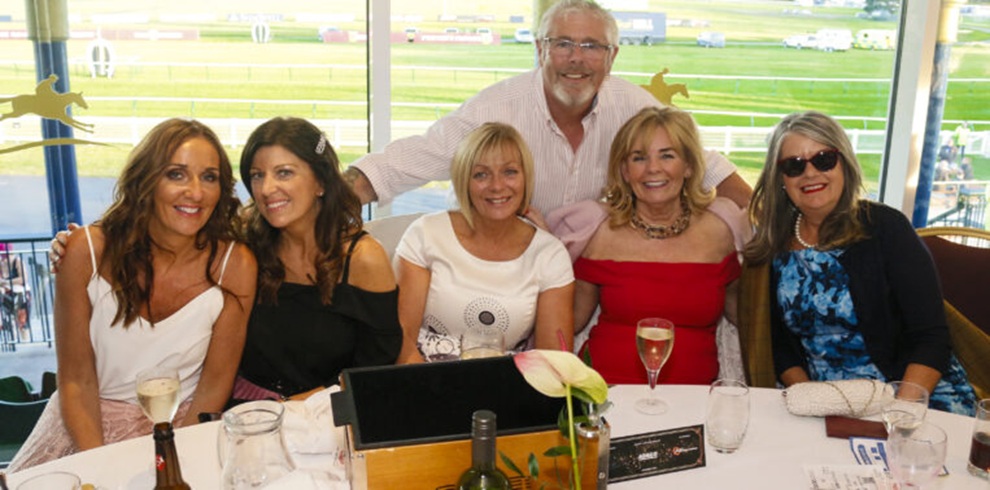 Six adults sit closely together around a table inside a venue overlooking a racetrack. The group poses facing the camera, with drinks on the table including wine glasses and bottles. One person stands behind the seated group with arms resting on the backs of chairs. The table holds menus, glassware, and a small floral decoration. Large windows in the background reveal a grassy racecourse and track rails in daylight.