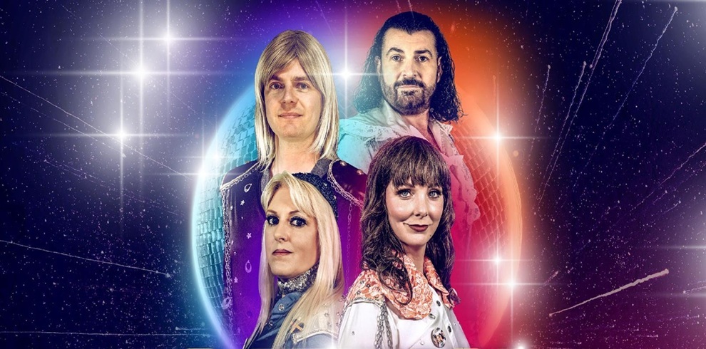 Promotional image featuring four performers arranged in two rows against a glittering disco-style background, with star-like light flares and a glowing circular backdrop in blue and purple tones. The performers wear retro-inspired costumes, creating a 1970s disco and tribute-show aesthetic.