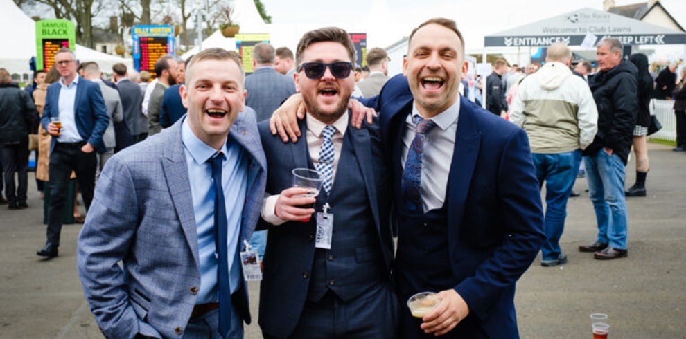 A photograph taken at an outdoor horse racing venue showing three people standing together in the foreground with arms around each other. All three are wearing suits and ties; the person on the left wears a light blue jacket, the person in the center wears a dark suit and sunglasses while holding a plastic cup, and the person on the right wears a navy suit and holds a drink. Behind them, a busy crowd of racegoers stands on paved ground near betting boards and signage, with trees and venue buildings visible in the background.