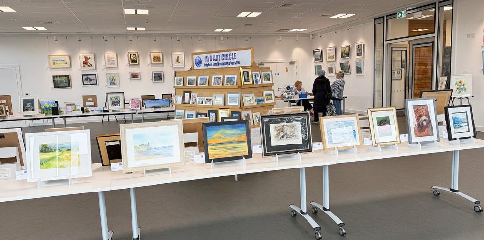 Interior view of the Ayr Art Circle Annual Exhibition at The Cutty Sark Centre. Dozens of framed artworks are displayed on tables, easels and gallery walls throughout a bright exhibition space. The collection includes landscapes, wildlife paintings, floral artwork and other original pieces. Visitors can be seen browsing the exhibition, with a banner reading "Ayr Art Circle Original Small Paintings for Sale" displayed towards the rear of the gallery.