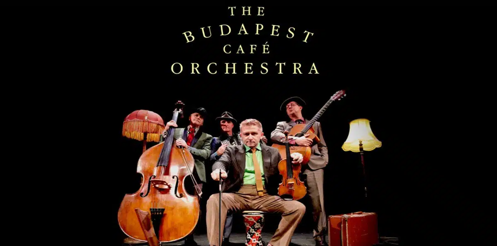 Promotional image for The Budapest Café Orchestra. Four musicians stand and sit on a dark stage surrounded by vintage-style props, including a large double bass, a violin, an acoustic guitar, two floor lamps, and a brown suitcase. One musician sits in the center holding a violin, while the others stand behind with their instruments. The band name, “THE BUDAPEST CAFÉ ORCHESTRA,” appears in large gold lettering above the group against a black background. The image is warmly lit, highlighting the instruments and performers.