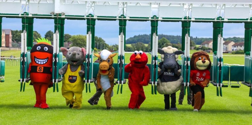 A wide photograph of a horse racing track starting gate on a grassy course. Seven people in large, colorful mascot costumes are walking forward from the open stalls beneath the white metal gate. The costumes include a red character with a large smiling face, a grey animal in yellow overalls, a brown animal with horns, a red fuzzy character, a sheep‑shaped costume with printed text on the front, and a lion‑like character. Trees, houses, and low hills are visible in the background under a bright sky.