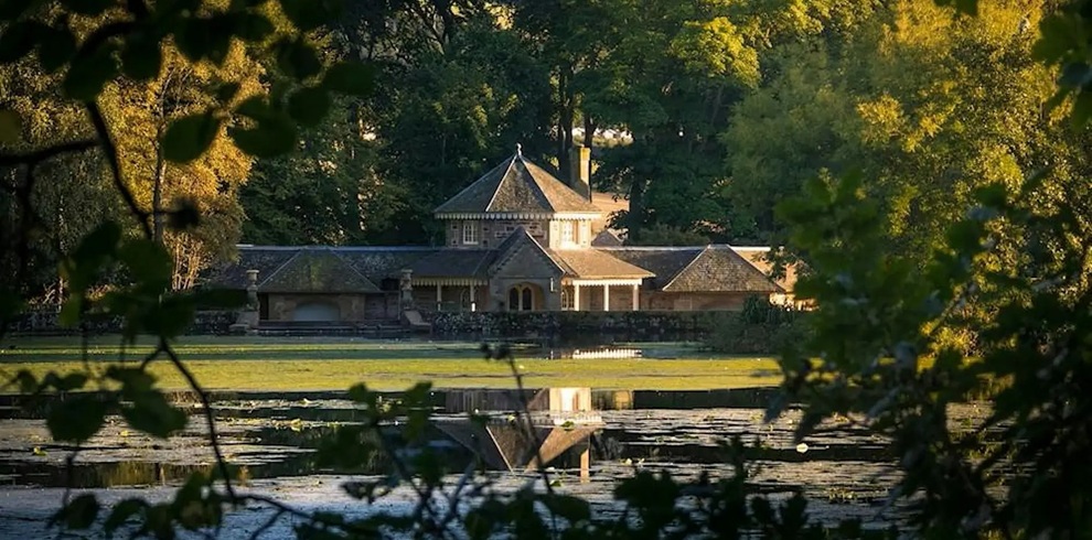 A tranquil view across a pond towards a stone pavilion-style building set within landscaped parkland, framed by leafy trees. The building and surrounding greenery are reflected in the still water in the foreground, with sunlight filtering through the foliage to create a calm, early-morning atmosphere.