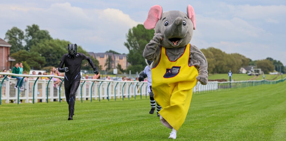 A wide photograph at a horse racing venue showing two people in mascot costumes running on a grassy racecourse. In the foreground, a grey elephant mascot wears bright yellow overalls with a logo on the chest, raising one arm while moving toward the camera. Behind and to the left, another mascot in a black full‑body costume is running in the same direction. White railings line the track, with spectators standing behind them and houses and trees visible in the background under a cloudy sky.