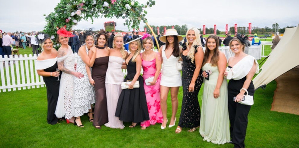 A group of people dressed in formal race-day outfits pose together on a grassy racecourse, standing beneath a decorative floral arch. They wear a mix of elegant dresses and headpieces, holding drinks, with crowds, track railings and festival flags visible in the background at a racecourse event.
