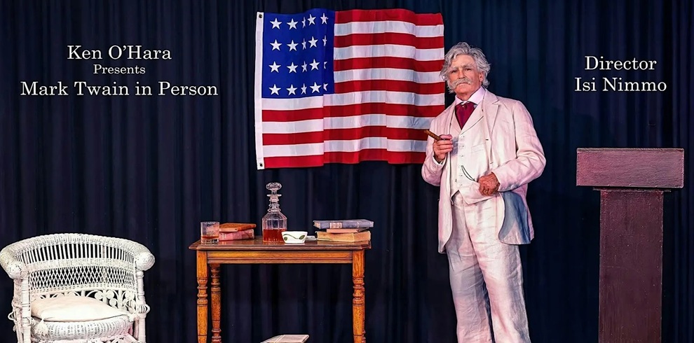 A staged theater scene shows a person in a light-colored vintage suit holding a cigar, standing beside a small wooden table with books, a glass decanter, and a cup. An American flag hangs against a dark curtain backdrop. A woven white chair sits to the left, and a wooden lectern stands to the right. Text on the image reads “Ken O’Hara Presents Mark Twain in Person” on the left and “Director Isi Nimmo” on the right.