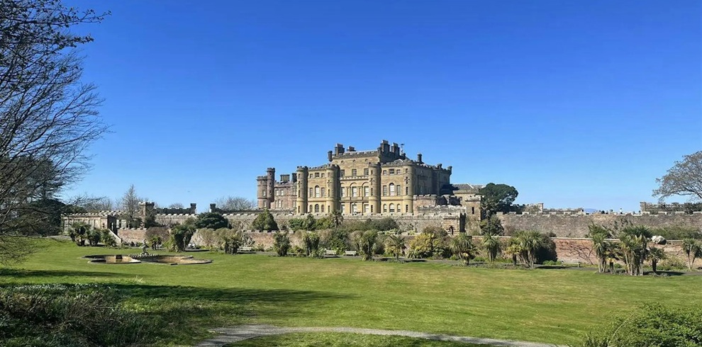 Wide view of a grand stone castle set behind a long outer wall, overlooking expansive green lawns and formal gardens under a clear blue sky. The castle stands centrally in the background, with trimmed grass, scattered trees, and garden features in the foreground, creating an open, parkland setting.