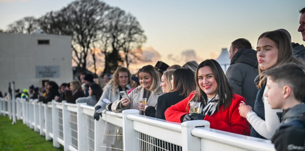 A wide photograph taken at an outdoor horse racing venue showing a line of spectators standing behind a white rail fence. Several people in coats and jackets lean on the rail, some holding clear plastic cups, while others look at mobile phones. One person in the foreground wears a bright red coat and stands near the center of the group. The crowd extends into the distance along the track, with grass to the left, trees and buildings in the background, and soft evening light in the sky.