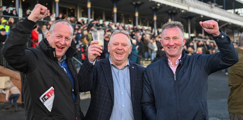 A photograph taken at an outdoor horse racing venue showing three people standing side by side in the foreground, facing the camera. Each person is wearing a dark jacket; two have one arm raised, and the person in the center is holding a clear plastic cup. Behind them, a large seated crowd fills a covered grandstand with metal railings and steps, creating a busy background under daylight conditions.