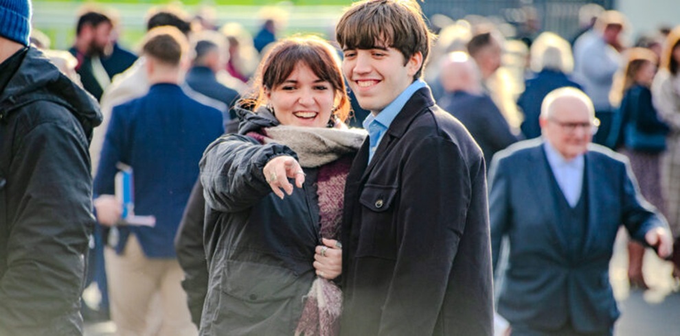 A photograph taken at an outdoor horse racing venue showing two people standing together in the foreground among a crowd of spectators. One person wears a dark jacket and scarf and is pointing forward, while the other person stands beside them wearing a dark coat and patterned shirt. Both are facing toward the camera area. Behind them, numerous racegoers move along a paved concourse, with railings, greenery, and figures in coats visible in soft daylight.