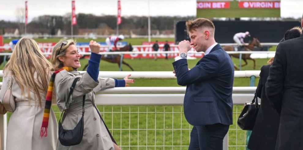 A photograph taken at a horse racing venue showing two people standing by a white rail fence in the foreground. One person on the left wears a light-colored coat and a striped scarf, raising one arm while turning toward the person opposite. The person on the right wears a navy suit and lifts an arm toward their face, with a watch visible on the wrist. Behind the rail, several racehorses with jockeys are running along the track. Other spectators stand nearby, and red signage reading “Virgin Bet” is visible in the distance under an overcast sky.