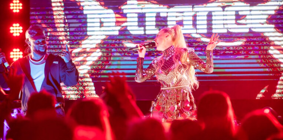 A photograph of a live music performance on an indoor stage viewed from the audience. Two performers stand under bright red and purple stage lighting; one performer at center right holds a microphone and wears a reflective, patterned outfit, while another performer to the left wears a dark jacket and also holds a microphone. A large LED screen behind them displays bold white lettering against a colorful abstract background. The silhouettes of audience members are visible in the foreground.