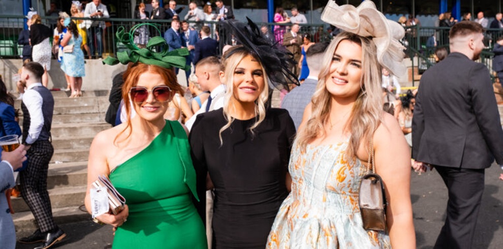A photograph taken at an outdoor horse racing venue showing three people standing side by side in the foreground. The person on the left is wearing a green one‑shoulder dress, sunglasses, and a decorative green headpiece, holding a small clutch bag. The person in the center is wearing a fitted black outfit with a black headpiece featuring layered fabric details. The person on the right is wearing a light‑colored patterned dress and a large cream‑colored fascinator, holding a small handbag. Behind them, a busy crowd of racegoers is visible near steps and railings, with people dressed in formal and semi‑formal attire under bright daylight.