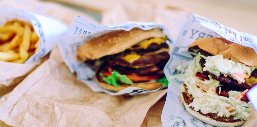 A market stall display showing rows of packaged baked goods, including steak pies, sausage rolls, pasties, and sliced pastries, arranged in red plastic crates and on a shelf above. Each item is sealed in clear plastic trays and labeled with blue-and-white stickers featuring prices and small Scottish flag designs.