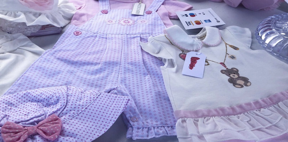A table display at a market showing pastel-colored baby clothes laid flat, including a pink-and-lilac patterned romper with snaps, a white dress with pink trim and a teddy bear illustration, and a matching polka-dot hat with a bow. Price tags and labels are attached, and other folded garments are partially visible around the display.