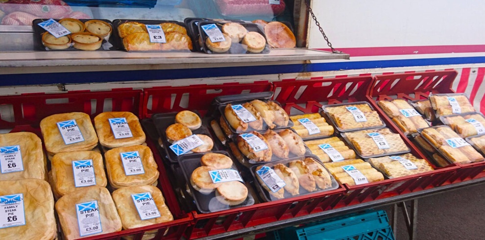 A market stall display with trays of packaged baked foods, including steak pies, sausage rolls, and pastries, arranged in red crates and on a shelf. Each item is sealed in clear plastic trays and labeled with blue-and-white stickers showing Scottish flags and prices.