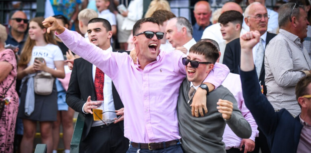 A photograph of a crowded outdoor grandstand at a horse racing event. In the foreground, two people wearing sunglasses stand close together with arms around each other; one person is wearing a light pink button‑down shirt and pointing toward the left of the frame, while the other is wearing a dark green top. Both have their mouths open and one arm raised. Around them, many spectators stand closely packed, some holding drinks, dressed in casual and semi‑formal clothing. Stadium seating and railings are visible in the background.