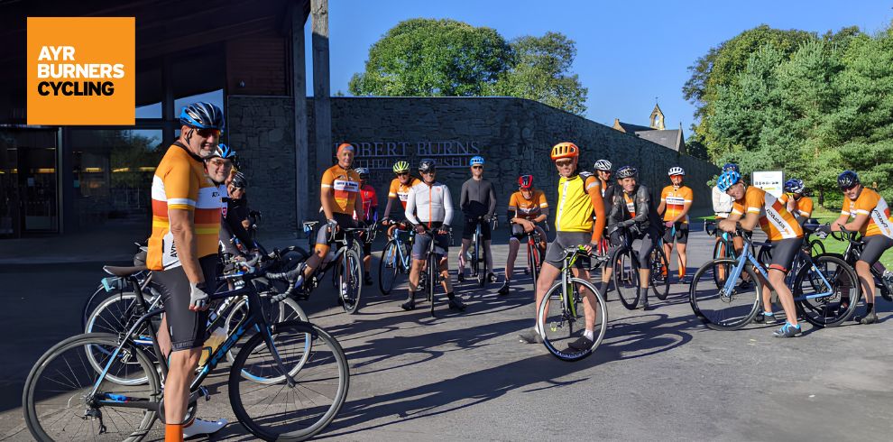 A large group of cyclists gathered outdoors in daylight, standing with their road bikes in front of a modern building with a stone wall. Many cyclists are wearing matching orange cycling jerseys, helmets, and cycling shorts, suggesting they are part of an organised club ride. The riders and bicycles are arranged loosely in a semi-circle, facing toward the camera. The bicycles have thin tyres and drop handlebars typical of road bikes. In the top-left corner of the image, there is a bright orange square graphic with white and black text that reads: “AYR BURNERS CYCLING.” Behind the group, the building features a stone wall with engraved text that includes the words “ROBERT BURNS BIRTHPLACE MUSEUM” (partially visible), indicating the location. The background includes tall green trees under a clear blue sky, and sunlight casts long shadows on the paved surface, suggesting the photo was taken in the morning or late afternoon. The overall scene conveys a group cycling meetup or event in a cultural or historic outdoor setting.