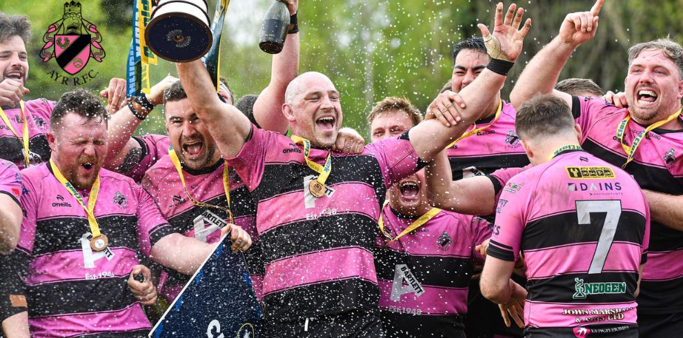 Ayr Rugby Football Club players in pink-and-black striped jerseys celebrate together, raising a trophy while champagne sprays into the air, with medals visible around their necks.