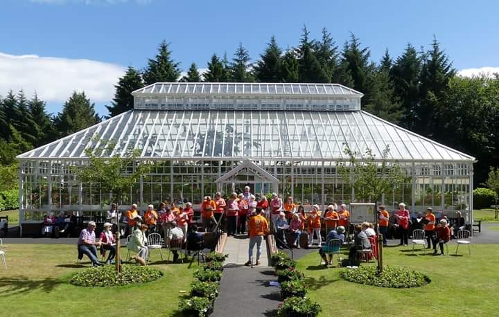An image of Belleisle Conservatory with a choir dressed in orange t-shirts singing in front of it.