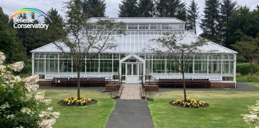 Promotional image for the Belleisle Conservatory. The image features an illustrated glass conservatory building surrounded by trees and landscaped gardens with flowerbeds in the foreground. The Belleisle Conservatory logo is in the top left-hand corner.