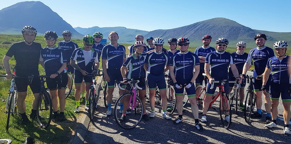 A group of around fifteen road cyclists stand together on a paved roadside, each holding or standing beside a bicycle. The cyclists wear matching navy-blue jerseys with white text reading “AYR ROADS CC,” along with helmets and cycling shorts. Several brightly colored bikes are visible, including pink, red, and black frames. The group is positioned in the foreground, arranged in a loose line facing the camera. Behind the cyclists, a scenic landscape stretches out with rolling green hills and a large, rounded mountain under clear blue skies. Bright sunlight casts strong shadows on the ground, emphasizing the crisp, clear weather and open countryside setting.