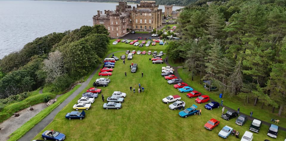 Aerial view of Culzean Castle grounds beside the coastline, showing rows of classic cars arranged on green lawns. The castle sits near the water with trees surrounding the area. Visitors are scattered among the cars, creating an organised display of various coloured vehicles.