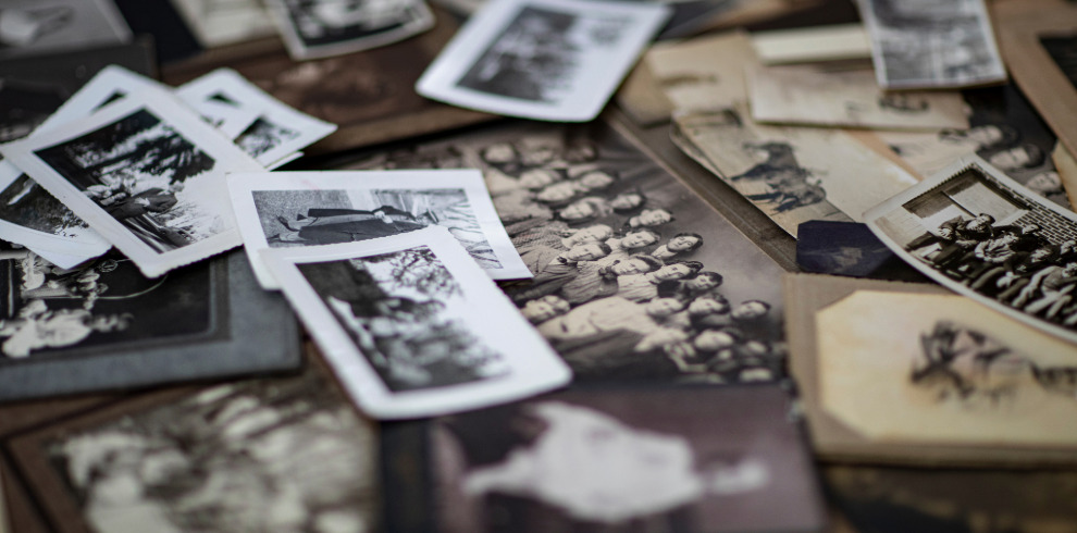 A close-up view of numerous vintage photographs scattered across a wooden surface. The images are mostly black-and-white or sepia-toned, showing groups of people, portraits, and outdoor scenes. Some photos overlap each other, while others are placed at slight angles. The lighting is soft and slightly dim, creating a nostalgic atmosphere, with the focus centered on a few prints in the foreground and the surrounding images gently blurred.
