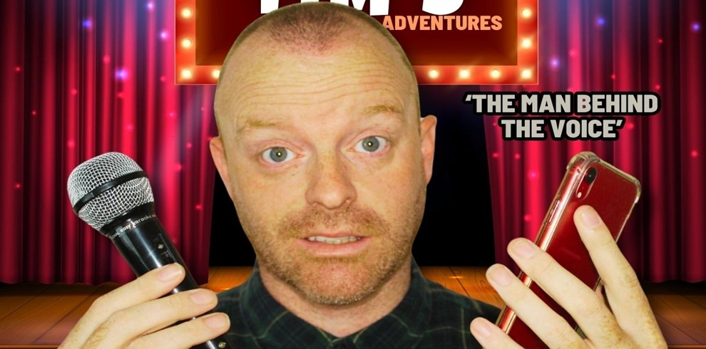 Close-up promotional image of a person standing on a stage set with red theatre curtains and warm marquee-style lights in the background. The person holds a handheld microphone in one hand and a red smartphone in the other, both raised near face level. A lit sign above shows partial text ending in “ADVENTURES.” To the right, bold text reads “The man behind the voice.” The stage floor is wooden, and the lighting creates a bright, performance-style atmosphere.