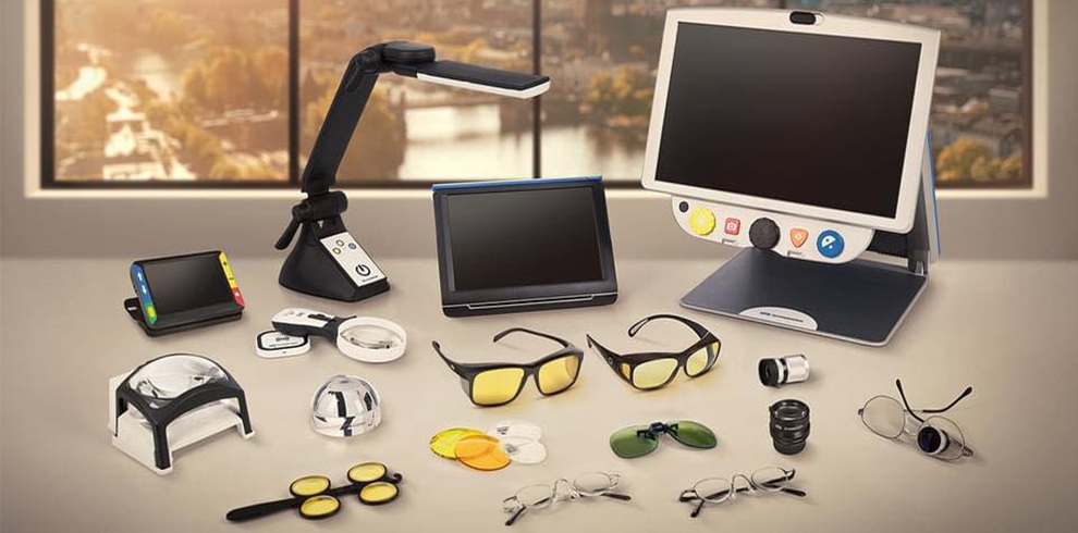 A display of assistive technology and visual aids for people with sight loss arranged on a table, including electronic magnifiers, a large screen reader device, task lighting, tinted glasses, handheld lenses, and various adaptive viewing tools, with a bright window view in the background.