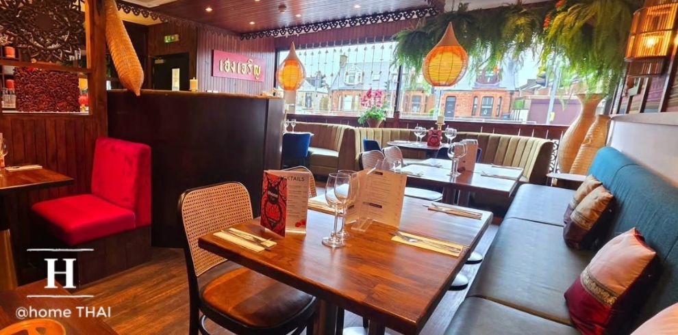 Interior of a Thai restaurant dining area with warm wooden décor. In the foreground, a wooden table is set with menus, wine glasses and cutlery. Surrounding seating includes cushioned bench seating and mixed chairs, with one red upholstered chair standing out. In the background, large windows let in natural light and show a street view of buildings outside. Hanging pendant lights and green foliage decor suspended from the ceiling add a cosy, ambient feel. A bar or service counter is visible to the left with wooden paneling. Text in the lower corner reads “@home THAI” with a stylised “H” logo.