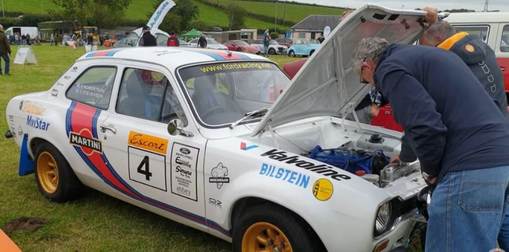 A white classic Ford Escort rally car parked on grass with its bonnet raised, exposing the engine. The car features racing decals and sponsor logos, including “Valvoline,” “Bilstein,” “Michelin,” and “Martin.” It has a large black number “4” on the door and stylized racing stripes in blue and red running along the side. The wheels are gold-colored. Two adults stand at the front of the car, leaning over the open engine bay, appearing to inspect or work on the engine. In the background, several other classic cars and tents are visible, along with people walking around the event area. The setting is an open grassy field under an overcast sky.