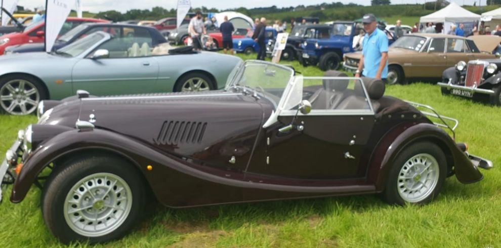 A dark burgundy or deep brown vintage-style convertible car parked on grass. The car has a long bonnet, rounded fenders, and a low windscreen, giving it a classic, retro design. The roof is down, revealing a simple interior. The wheels are silver with a multi-spoke design. Behind the convertible, multiple other classic cars are parked, including a green convertible, a blue vehicle, and a gold or bronze car. White event tents and flags are visible in the background, along with a number of people walking and viewing the cars. The setting is a grassy outdoor event area with rolling green hills in the distance and soft daylight.