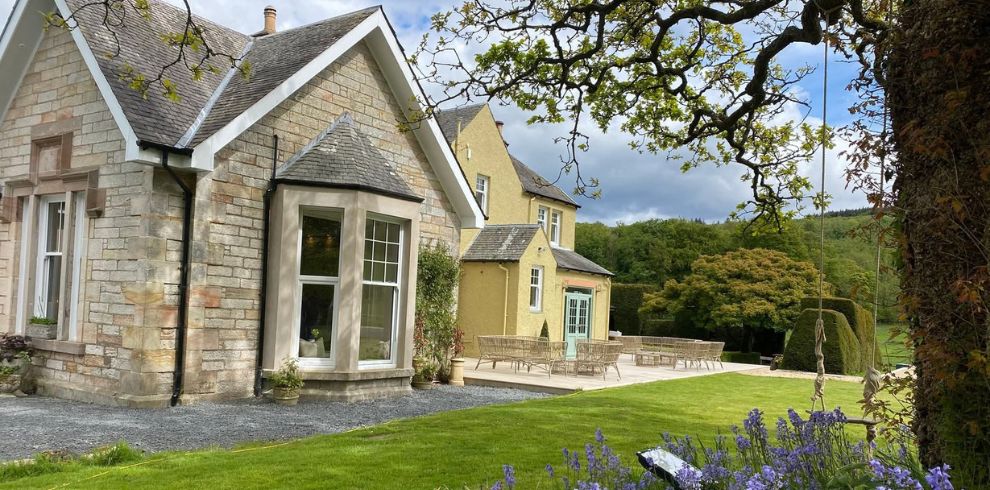 Exterior of Ladyburn House Country Retreat, Scotland An outdoor photograph of a countryside property set within landscaped grounds. Foreground: A well-maintained lawn with green grass and clusters of purple flowers near the bottom edge. Right side foreground: A large tree with twisting branches and textured bark extends into the frame. A rope swing hangs from one branch. Centre: A paved patio area with outdoor wooden or wicker seating arranged for relaxation. Building: A light stone house with a pitched slate roof and large bay window faces the garden. Adjacent is a pale yellow extension with multiple doors and windows. Background: Rolling greenery and trees suggest a rural or hillside setting. Sky: Partly cloudy with patches of blue and sunlight. The overall scene conveys a tranquil countryside retreat with spacious outdoor living areas and charming architectural features.