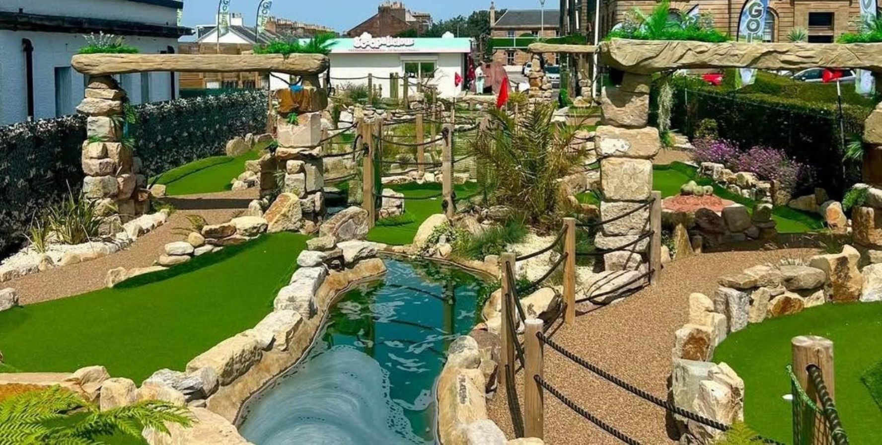 Adventure-themed mini golf course featuring artificial green turf, stone archways, rock-lined paths, rope railings, and a narrow water feature running through the center. Landscaping includes boulders, shrubs, and palm-style plants, with buildings visible in the background on a sunny day.