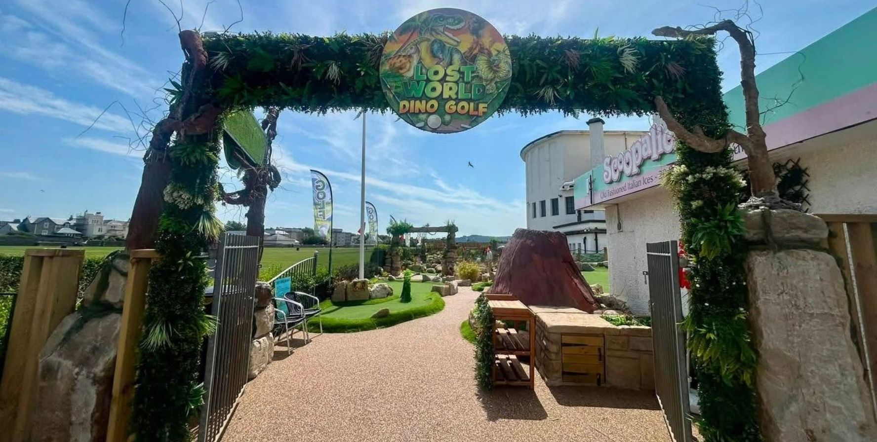 Entrance to Lost World Dino Golf, marked by a large decorative arch covered in greenery and a circular sign reading “Lost World Dino Golf.” A winding pathway leads into an adventure golf course with artificial turf, rock features, and dinosaur-themed landscaping beside a building under a bright blue sky.