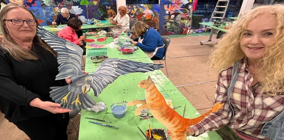 Two people in the foreground sit at a long, green-covered table in an indoor art space. Each person holds a painted cutout—one of a gray bird in flight with wings spread, and the other of an orange cat mid-stride. Paintbrushes, cups of water, and art supplies are scattered across the table. In the background, several other people sit and paint at the same table. The walls behind them are covered with colorful, detailed murals featuring animals and abstract designs, adding a vibrant, creative atmosphere to the room.